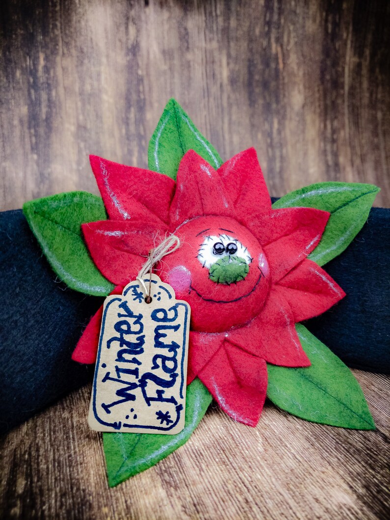 May include: A handmade felt poinsettia flower with green leaves and a smiling face. A small tag with the words "Winter Flame" is attached. The flower is red with white stitching and pink cheeks.