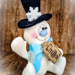 Puede incluir: Un mu&ntilde;eco de nieve blanco con un sombrero de copa negro, un lazo azul y una etiqueta marr&oacute;n que dice "Chillin'"
