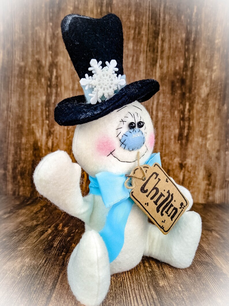 Puede incluir: Un mu&ntilde;eco de nieve blanco con un sombrero de copa negro, un lazo azul y una etiqueta marr&oacute;n que dice "Chillin'"