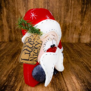 May include: A fabric Santa Claus ornament featuring a red hat with white star embroidery, a white beard, and a small green pine sprig. A tan cardboard tag is attached with twine, featuring the handwritten text Little Nicky in black ink.