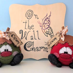 May include: Two felt pumpkins with stitched details, one green and one red, with googly eyes and black shoes. Each pumpkin has a brown tag with the words "Ooey" and "Gooey" written on them. The pumpkins are sitting in front of a wooden sign with the words "On the Wall Charm" written on it.