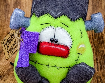 May include: A green felt Frankenstein monster with black felt hair, stitched seams, and a red mouth. The monster has two black eyes and a purple felt scarf. The monster is sitting on a wooden surface with a tag that says "Freaky Frank".