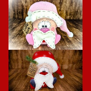 May include: Two plush gnome dolls with stitched details, one in pink and one in red. Each features a floppy hat with white snowflake embroidery, a large white moustache, and a tan tag attached to the side that reads Little Nicky in black ink.