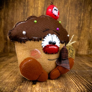May include: A brown felt cupcake with a red felt cherry on top. The cupcake has a white felt face with red felt lips and black felt eyes. The cupcake is holding a small brown felt tag that says "Sugar Rush".
