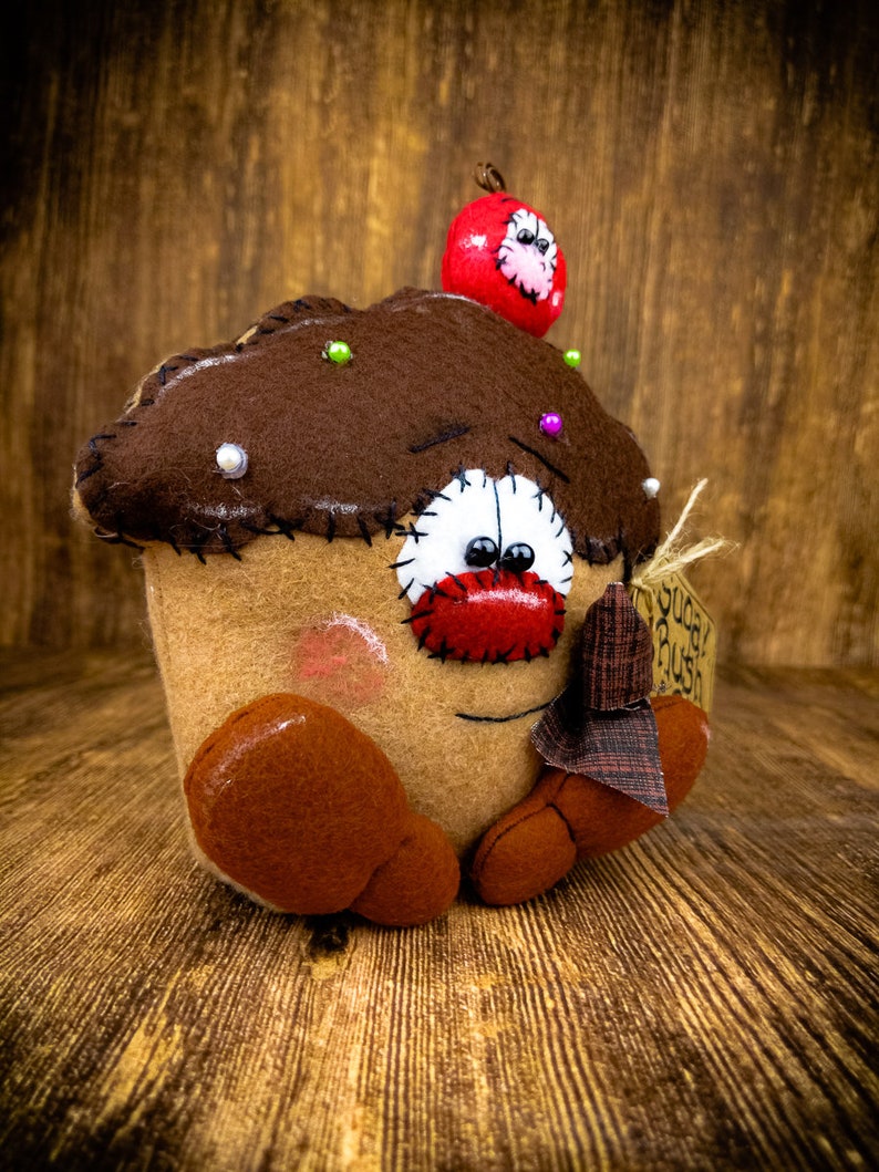 May include: A brown felt cupcake with a red felt cherry on top. The cupcake has a white felt face with red felt lips and black felt eyes. The cupcake is holding a small brown felt tag that says "Sugar Rush".
