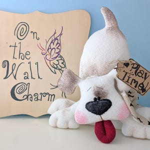 May include: A white felt dog with brown ears and a black nose, sticking its tongue out. The dog is holding a brown tag that says "Play Time". The dog is sitting in front of a wooden sign that says "On the Wall Charm" with a butterfly design.