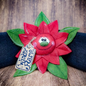 May include: A felt poinsettia flower with a smiling face and green leaves. The flower is red with a tag that says "Winter Flame". The flower is placed on a black felt surface, all on a wooden background.