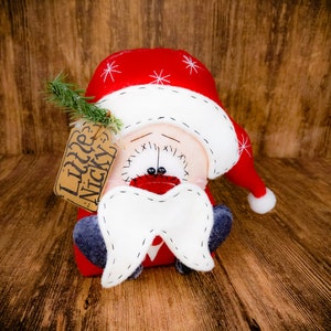 May include: A plush Santa Claus doll wearing a red hat with white snowflake embroidery and a white pom-pom. The doll features a large white felt moustache, dark grey feet, and a brown paper tag attached with green pine needles reading Little Nicky.