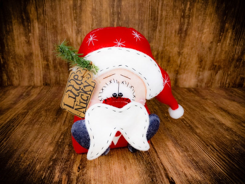 May include: A plush Santa Claus doll wearing a red hat with white snowflake embroidery and a white pom-pom. The doll features a large white felt moustache, dark grey feet, and a brown paper tag attached with green pine needles reading Little Nicky.