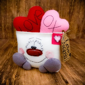 May include: A Valentine's Day themed plush toy with a white container featuring a stitched face and gray feet. Two heart-shaped pillows, one red with "LOVE" and one pink with "XO", sit inside. A tag reads "Love Notes".
