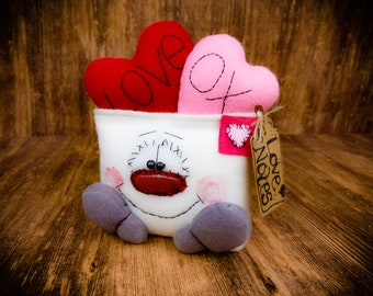 Puede incluir: Un peluche con temática de San Valentín con un recipiente blanco con una cara cosida y pies grises. Dos almohadas en forma de corazón, una roja con "LOVE" y otra rosa con "XO", están dentro. Una etiqueta dice "Love Notes".
