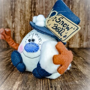May include: A white felt snowman with a blue hat, a blue scarf, and a brown felt tag that says "Snow Ball".