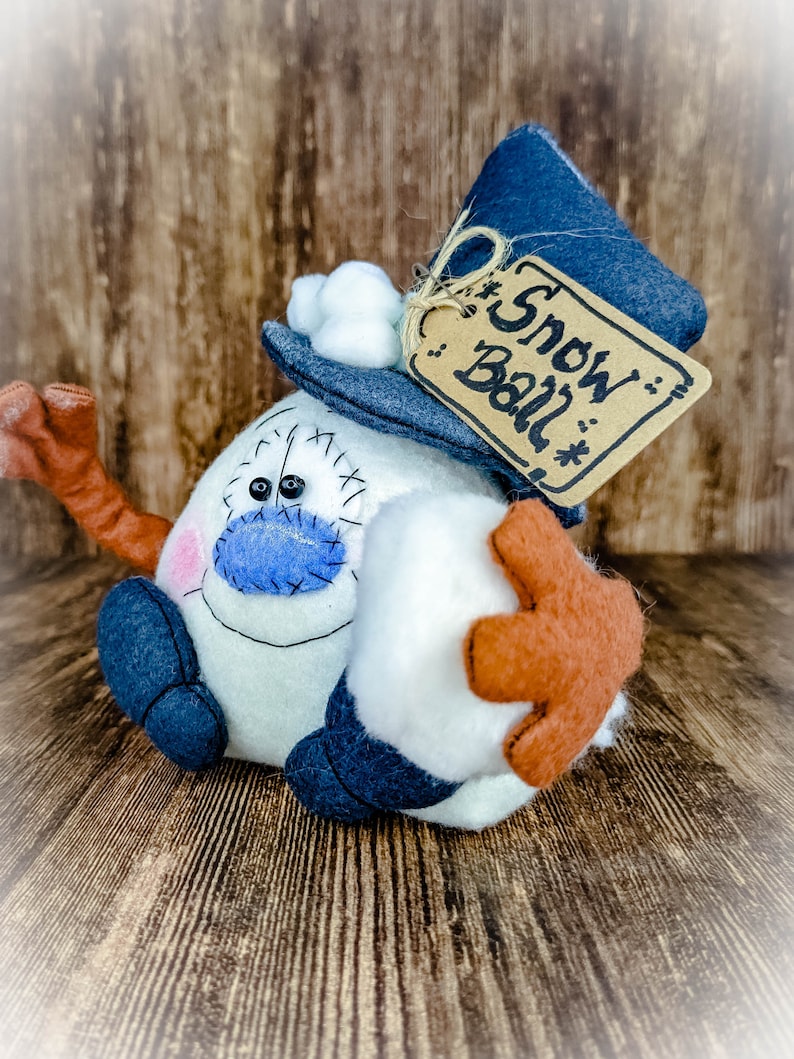 May include: A white felt snowman with a blue hat, a blue scarf, and a brown felt tag that says "Snow Ball".