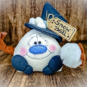 May include: A white snowman with a blue nose and a blue hat with a white pom-pom. The snowman is holding a white snowball in its right hand. The snowman has a brown arm and a tag that says "Snow Ball" hanging from its hat.