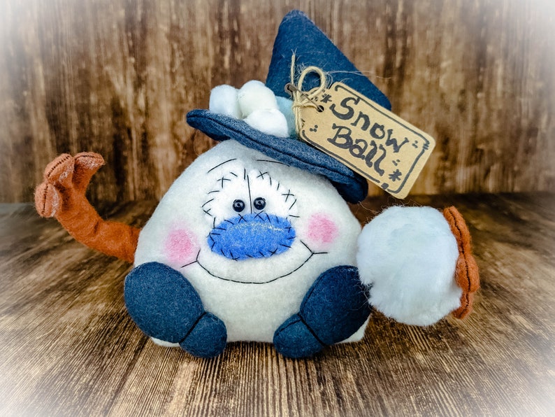 May include: A white snowman with a blue nose and a blue hat with a white pom-pom. The snowman is holding a white snowball in its right hand. The snowman has a brown arm and a tag that says "Snow Ball" hanging from its hat.