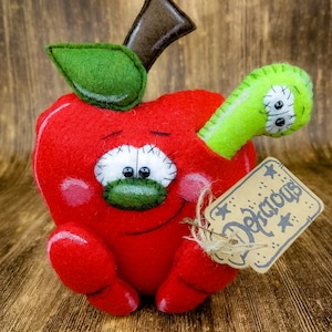 May include: A red felt apple with a green felt leaf and a brown felt stem. The apple has a smiling face with black eyes and a green felt mouth. A green felt worm with black eyes is sticking out of the apple. The apple is holding a brown tag that says "Delicious" in black lettering.