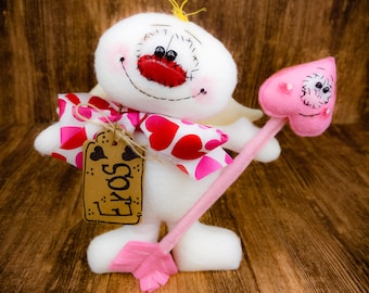 Puede incluir: Un ángel de fieltro blanco con una flecha rosa en forma de corazón y una etiqueta que dice "Love". El ángel tiene una boca roja y mejillas rosadas. El ángel sostiene la flecha en su mano derecha.