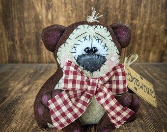 Può includere: Un orso di stoffa patchwork marrone e beige con un fiocco gingham rosso e bianco. L'orso ha un viso cucito con occhi neri e un naso nero. L'orso è seduto su una superficie di legno marrone e ha un'etichetta che dice "Brownie".