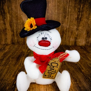 Puede incluir: Un mu&ntilde;eco de nieve blanco con un sombrero de copa negro y un lazo rojo. El mu&ntilde;eco de nieve tiene una cara de reloj con n&uacute;meros negros y una nariz roja. El mu&ntilde;eco de nieve sostiene una etiqueta marr&oacute;n que dice "Cheers".