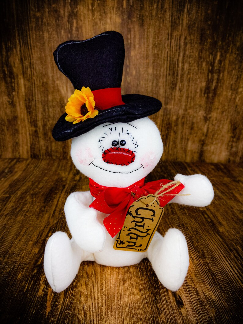 Puede incluir: Un mu&ntilde;eco de nieve blanco con un sombrero de copa negro y un lazo rojo. El mu&ntilde;eco de nieve tiene una cara de reloj con n&uacute;meros negros y una nariz roja. El mu&ntilde;eco de nieve sostiene una etiqueta marr&oacute;n que dice "Cheers".