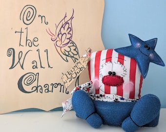 May include: A handmade patriotic doll with red, white, and blue fabric. The doll is wearing a blue star on its head and has a red, white, and blue striped shirt. The doll is sitting on a white surface with a sign that says "On the Wall Charms" and a tag that says "Happy 4th of July".