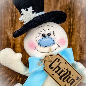 Puede incluir: Un mu&ntilde;eco de nieve blanco con un sombrero de copa negro, un lazo azul y una etiqueta marr&oacute;n que dice "Chillin".