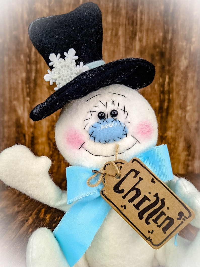 Puede incluir: Un mu&ntilde;eco de nieve blanco con un sombrero de copa negro, un lazo azul y una etiqueta marr&oacute;n que dice "Chillin".