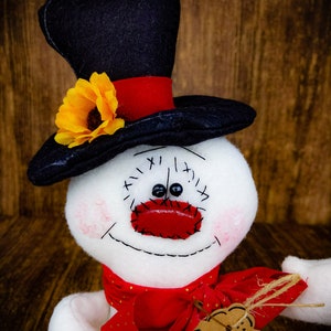 Puede incluir: Un mu&ntilde;eco de nieve blanco con un sombrero de copa negro, una bufanda roja y una nariz roja. El mu&ntilde;eco de nieve tiene una sonrisa cosida y ojos negros. El sombrero tiene un girasol amarillo en el lateral. El mu&ntilde;eco de nieve tiene una etiqueta marr&oacute;n que dice "Regalo" en ella.