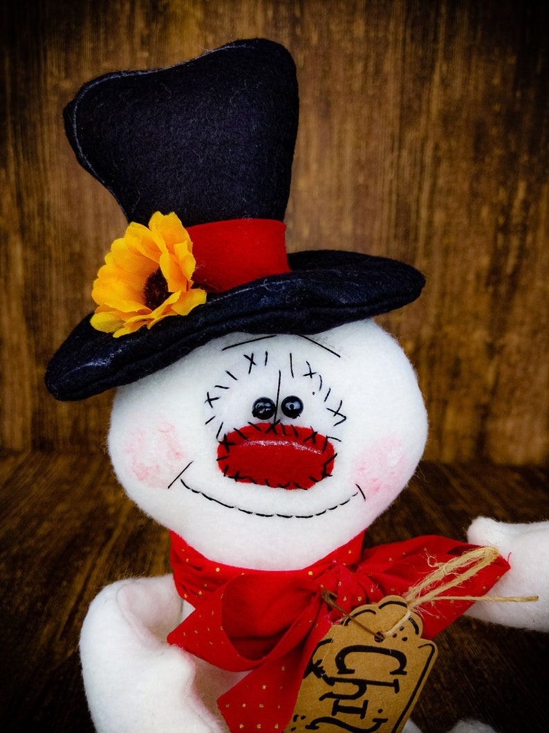 Puede incluir: Un mu&ntilde;eco de nieve blanco con un sombrero de copa negro, una bufanda roja y una nariz roja. El mu&ntilde;eco de nieve tiene una sonrisa cosida y ojos negros. El sombrero tiene un girasol amarillo en el lateral. El mu&ntilde;eco de nieve tiene una etiqueta marr&oacute;n que dice "Regalo" en ella.