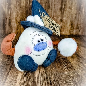 May include: A white snowman with a blue nose and black eyes wearing a blue hat with a white pom-pom and a brown tag that says "Snow Ball".