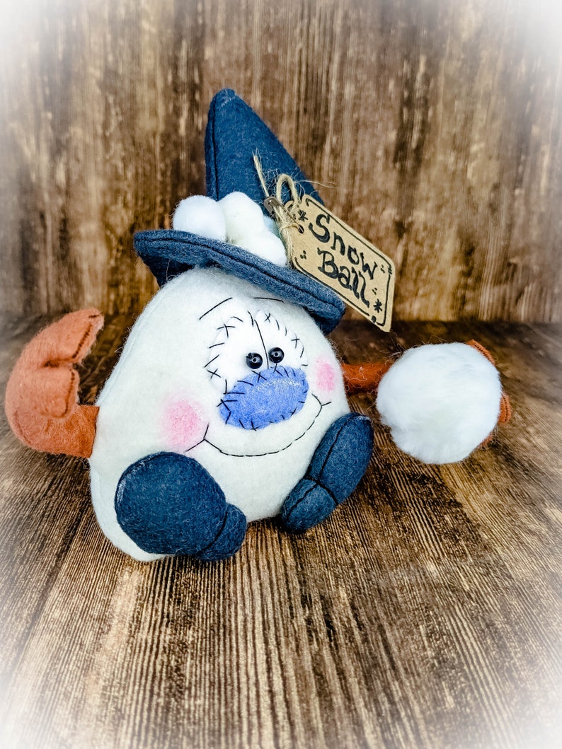 May include: A white snowman with a blue nose and black eyes wearing a blue hat with a white pom-pom and a brown tag that says "Snow Ball".