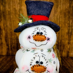 Pu&ograve; includere: Un pupazzo di neve fatto di tre sfere di tessuto bianco con un cappello a cilindro nero, un nastro rosso e campanelli dorati. Il pupazzo di neve ha un sorriso cucito e un naso di feltro arancione. Il pupazzo di neve indossa una sciarpa di tessuto bianco con un motivo verde e bianco.