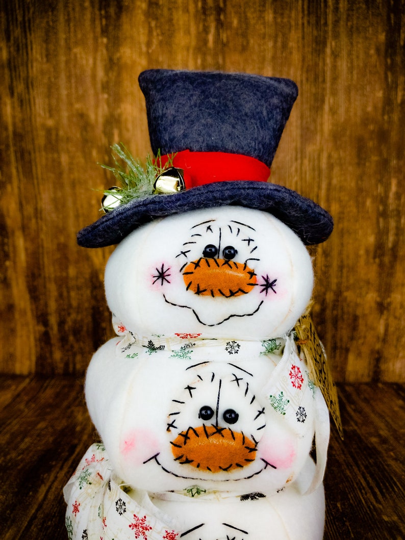 Pu&ograve; includere: Un pupazzo di neve fatto di tre sfere di tessuto bianco con un cappello a cilindro nero, un nastro rosso e campanelli dorati. Il pupazzo di neve ha un sorriso cucito e un naso di feltro arancione. Il pupazzo di neve indossa una sciarpa di tessuto bianco con un motivo verde e bianco.