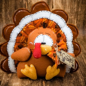 May include: A brown and orange felt turkey with a white felt bib and a tag that says "Giblet". The turkey is sitting on a wooden surface.