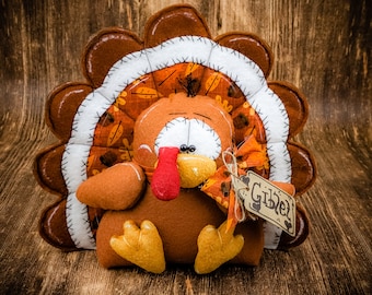 Puede incluir: Un pavo de fieltro marrón y naranja con un babero de fieltro blanco y una etiqueta que dice "Giblet". El pavo está sentado sobre una superficie de madera.