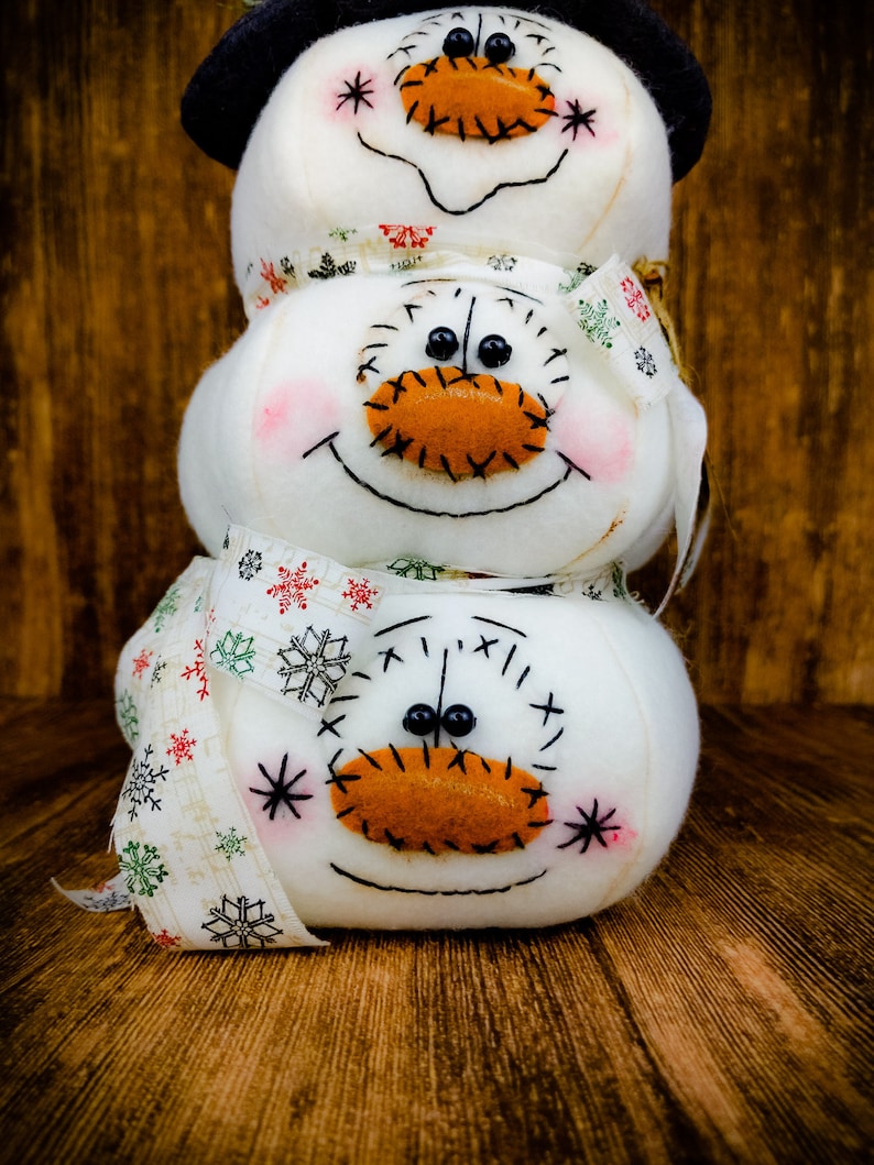 Pu&ograve; includere: Un pupazzo di neve fatto di tre sfere di tessuto bianco con occhi, nasi e bocche cuciti. Il pupazzo di neve indossa una sciarpa a motivi bianchi e verdi e un cappello nero in cima.