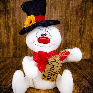 Puede incluir: Un mu&ntilde;eco de nieve blanco con un sombrero de copa negro, un lazo rojo y una etiqueta marr&oacute;n que dice "Chilly".