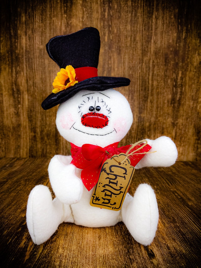 Puede incluir: Un mu&ntilde;eco de nieve blanco con un sombrero de copa negro, un lazo rojo y una etiqueta marr&oacute;n que dice "Chilly".