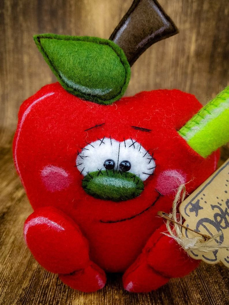 May include: A red felt apple with a green felt leaf and stem, a green felt pencil sticking out of the side, and a brown tag with the word "Delic" on it.