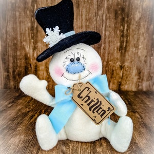 Puede incluir: Un mu&ntilde;eco de nieve blanco con un sombrero de copa negro, un lazo azul y una etiqueta marr&oacute;n que dice "Chillin".