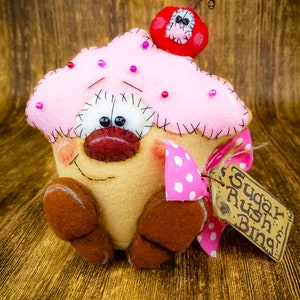 May include: A pink and brown felt cupcake with a red felt cherry on top. The cupcake has a smiling face with black eyes and a pink nose. The cupcake is wearing a pink and white polka dot bow and has a brown felt tag that says "Sugar Rush Bing!"