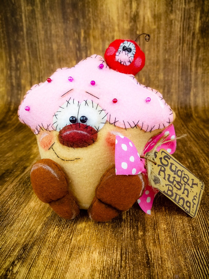 May include: A pink and brown felt cupcake with a red felt cherry on top. The cupcake has a smiling face with black eyes and a pink nose. The cupcake is wearing a pink and white polka dot bow and has a brown felt tag that says "Sugar Rush Bing!"