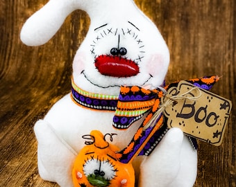 May include: White felt ghost with a red mouth, black eyes, and a purple and green ribbon around its neck. The ghost is holding a small orange felt pumpkin with a green eye and black mouth. A brown tag with the word "Boo" is attached to the ribbon.
