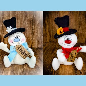 Puede incluir: Dos figuras de mu&ntilde;eco de nieve blancas con sombreros negros, una con un lazo azul y la otra con un lazo rojo. Ambos mu&ntilde;ecos de nieve tienen una etiqueta marr&oacute;n que dice "Chillin".
