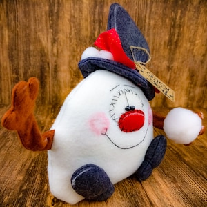 May include: A white snowman plush toy with a blue hat, red nose, and black eyes. The snowman is holding a white snowball in its right hand. The snowman is sitting on a wooden surface.