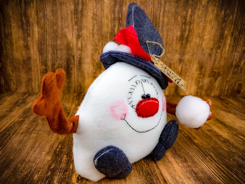 May include: A white snowman plush toy with a blue hat, red nose, and black eyes. The snowman is holding a white snowball in its right hand. The snowman is sitting on a wooden surface.