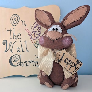 May include: A brown felt bunny with a beige bow and a tag that says "loppy". The bunny is sitting on a white surface in front of a wooden sign that says "On the Wall Charm".