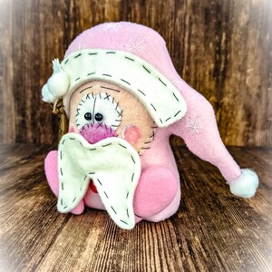 May include: A handcrafted felt doll featuring a soft pink pointed hat with white embroidered snowflake details and a white pom-pom. The doll has a cream-coloured face with stitched black eyes, a pink nose, and a large, cream-coloured felt moustache.