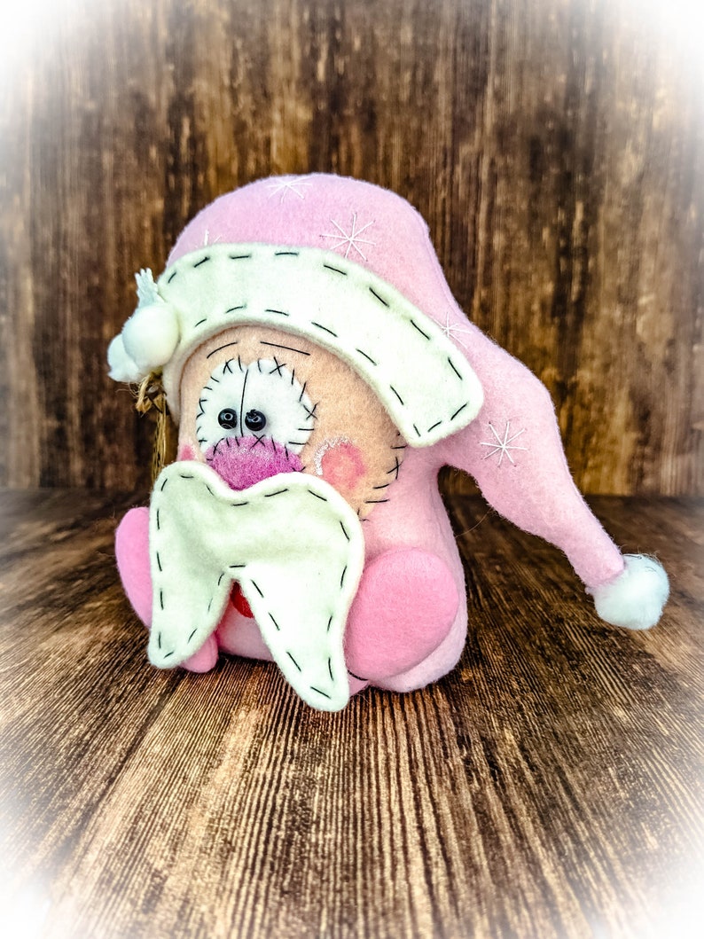 May include: A handcrafted felt doll featuring a soft pink pointed hat with white embroidered snowflake details and a white pom-pom. The doll has a cream-coloured face with stitched black eyes, a pink nose, and a large, cream-coloured felt moustache.