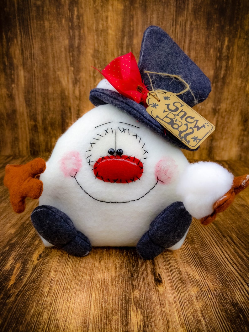 May include: A white snowman with a red nose and black eyes, wearing a blue hat with a red bow and a brown tag that says "Snow Ball".